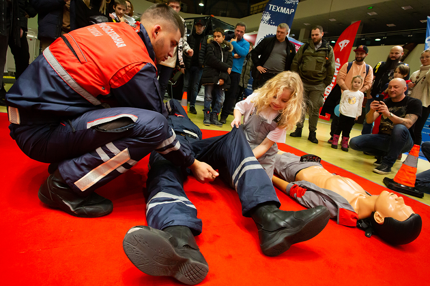 02.12.2022, Москва, Выставка, посвященная путешествиям и технике. «Поехали 2022» в Экспоцентре. Имитация с помощью манекена оказания первой медицинской помощи человеку, попавшему в аварию. Маленькая девочка помогает спасателям.
